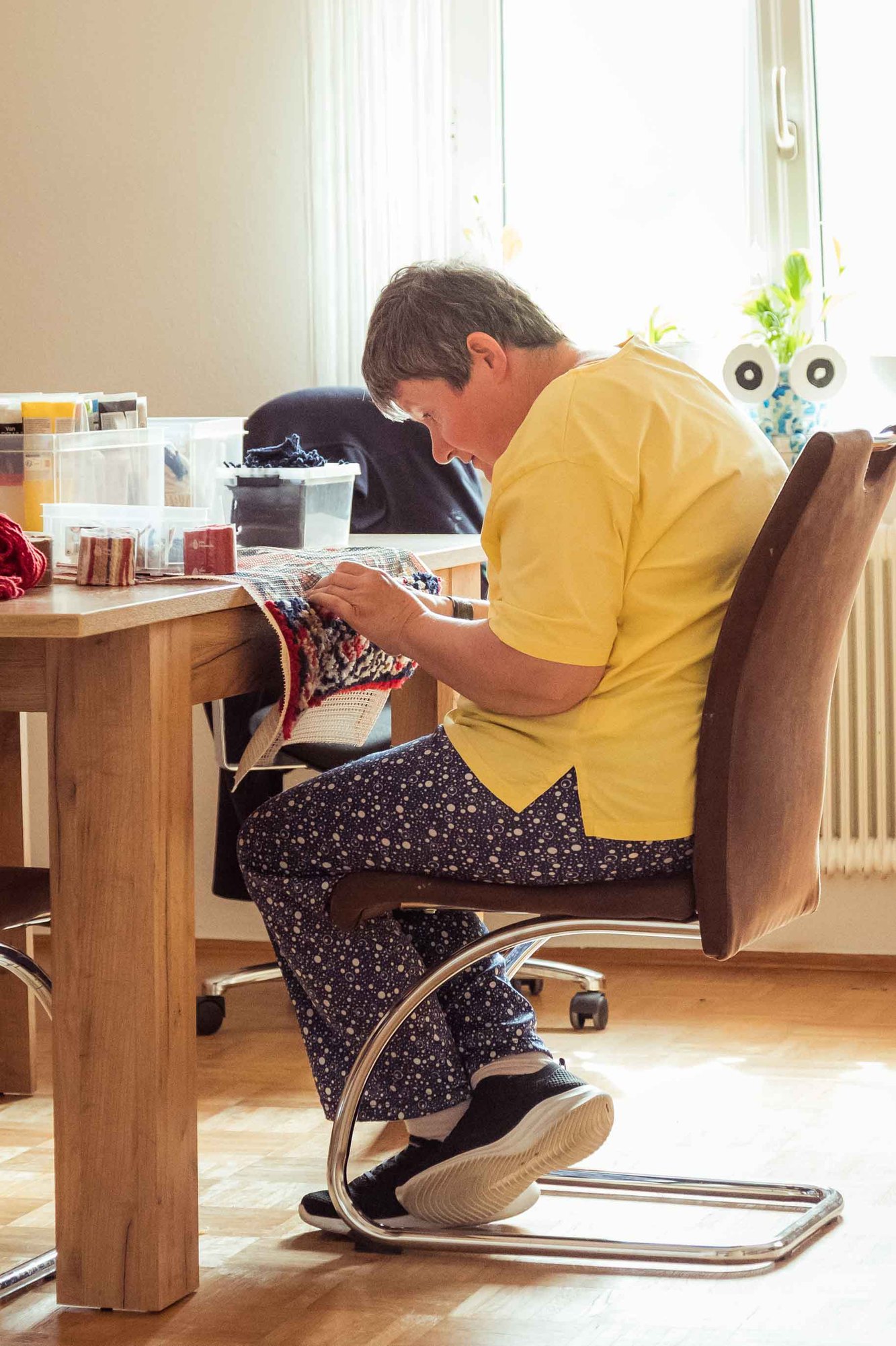 Eine Frau stickt konzentriert an einem Tisch am Fenster